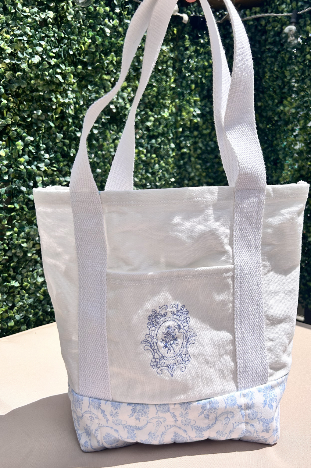 A white canvas tote bag with a Blue floral pattern on the lower part and a Blue crest embroidered design on the front pocket. 100% Canvas cotton