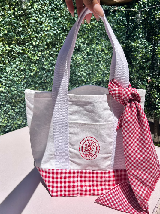 A white canvas tote bag with a red gingham pattern on the lower part and a red embroidered design on the front pocket. Quilted square base to hold heavy items