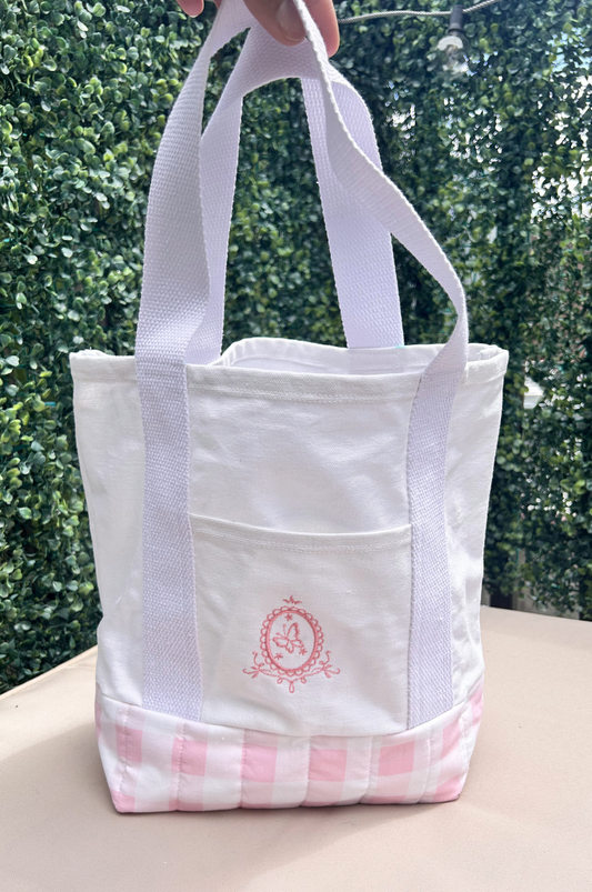 A white canvas tote bag with  a pink gingham pattern on the lower part and a pink butterfly embroidered design on the front pocket. 100% canvas cotton