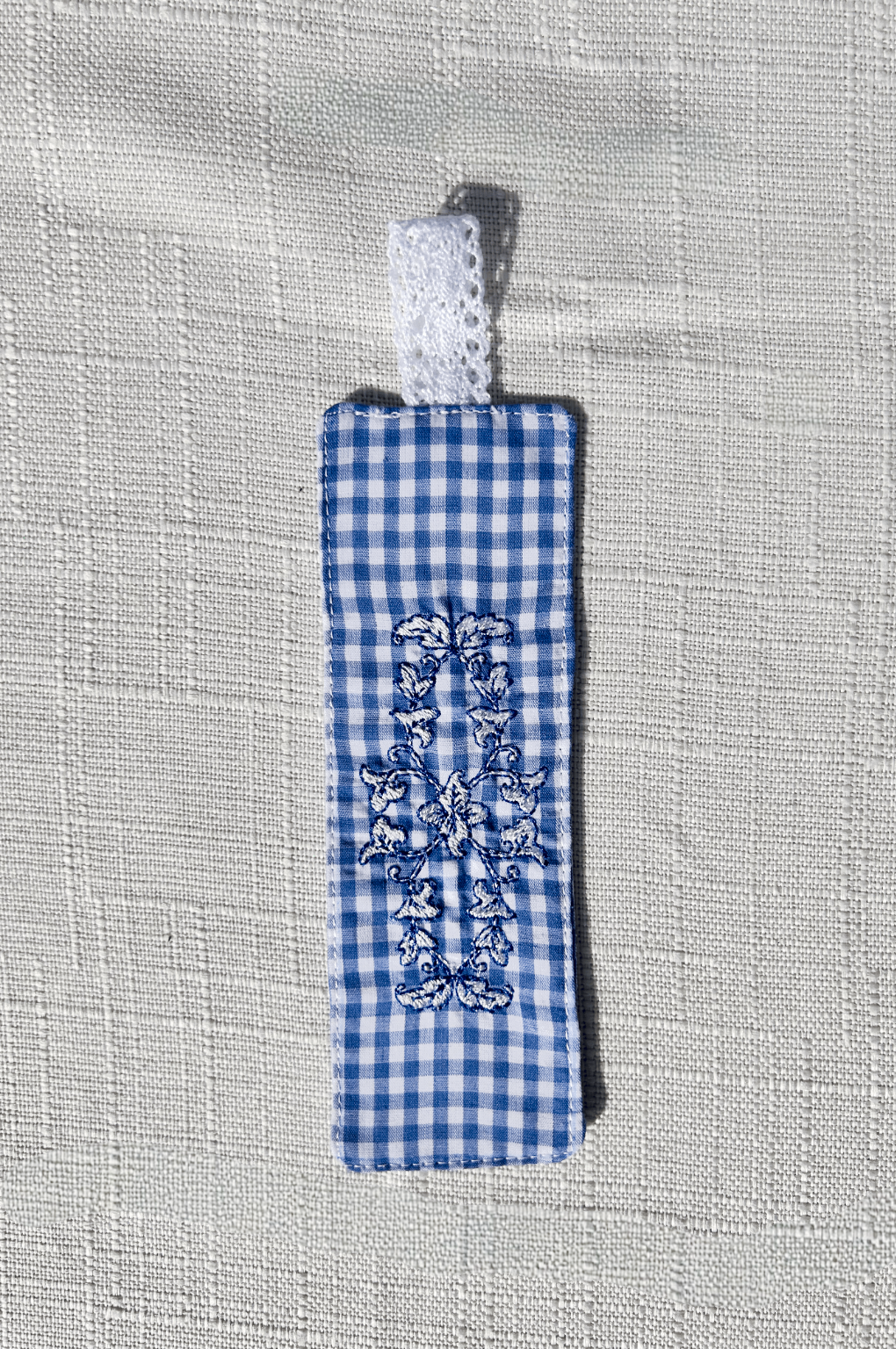 Handmade blue and white gingham fabric bookmark with an embroidered floral crest design and a white eyelet lace loop at the top.