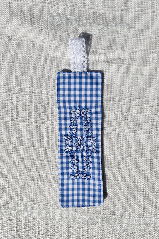 Handmade blue and white gingham fabric bookmark with an embroidered floral crest design and a white eyelet lace loop at the top.